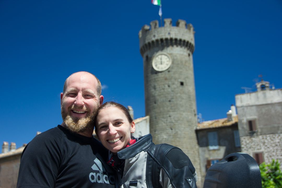 Smiling couple posing in front of a stone clock tower under a bright blue sky
