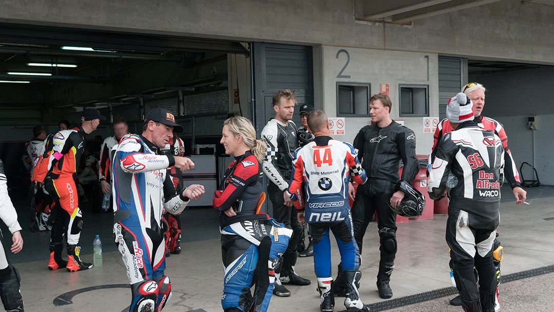 Motorcycle racers in colorful suits chatting in a pit garage, with bikes and crew in the background