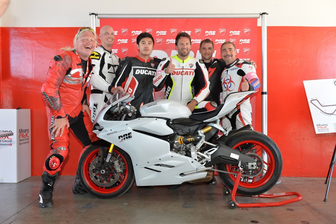 Five motorcycle racers posing with a white sport bike in a red garage or team area