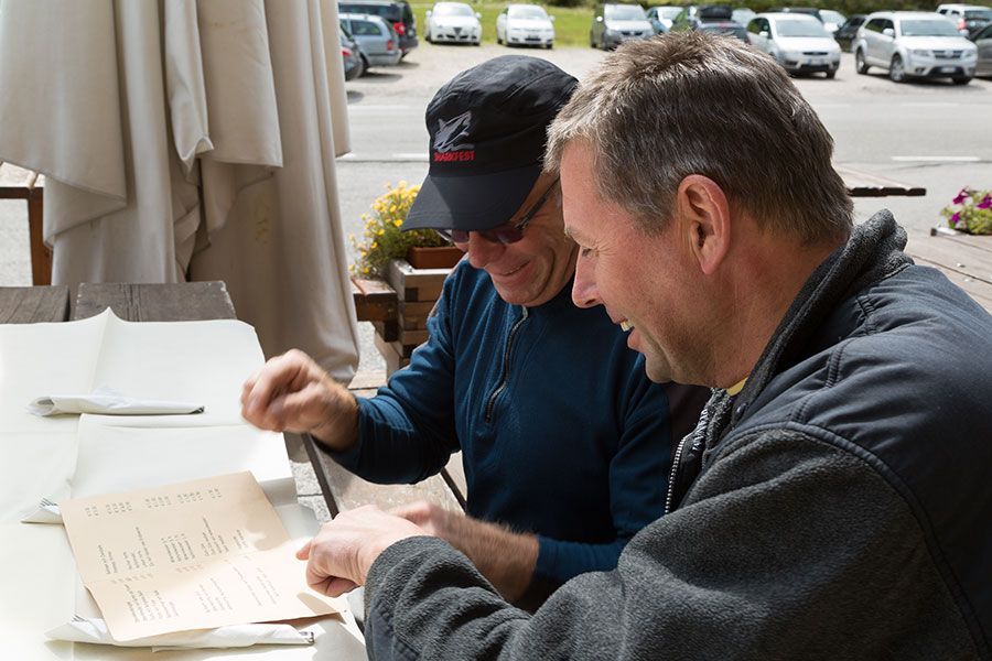 Two men reviewing papers at an outdoor cafe table, with parked cars in the background