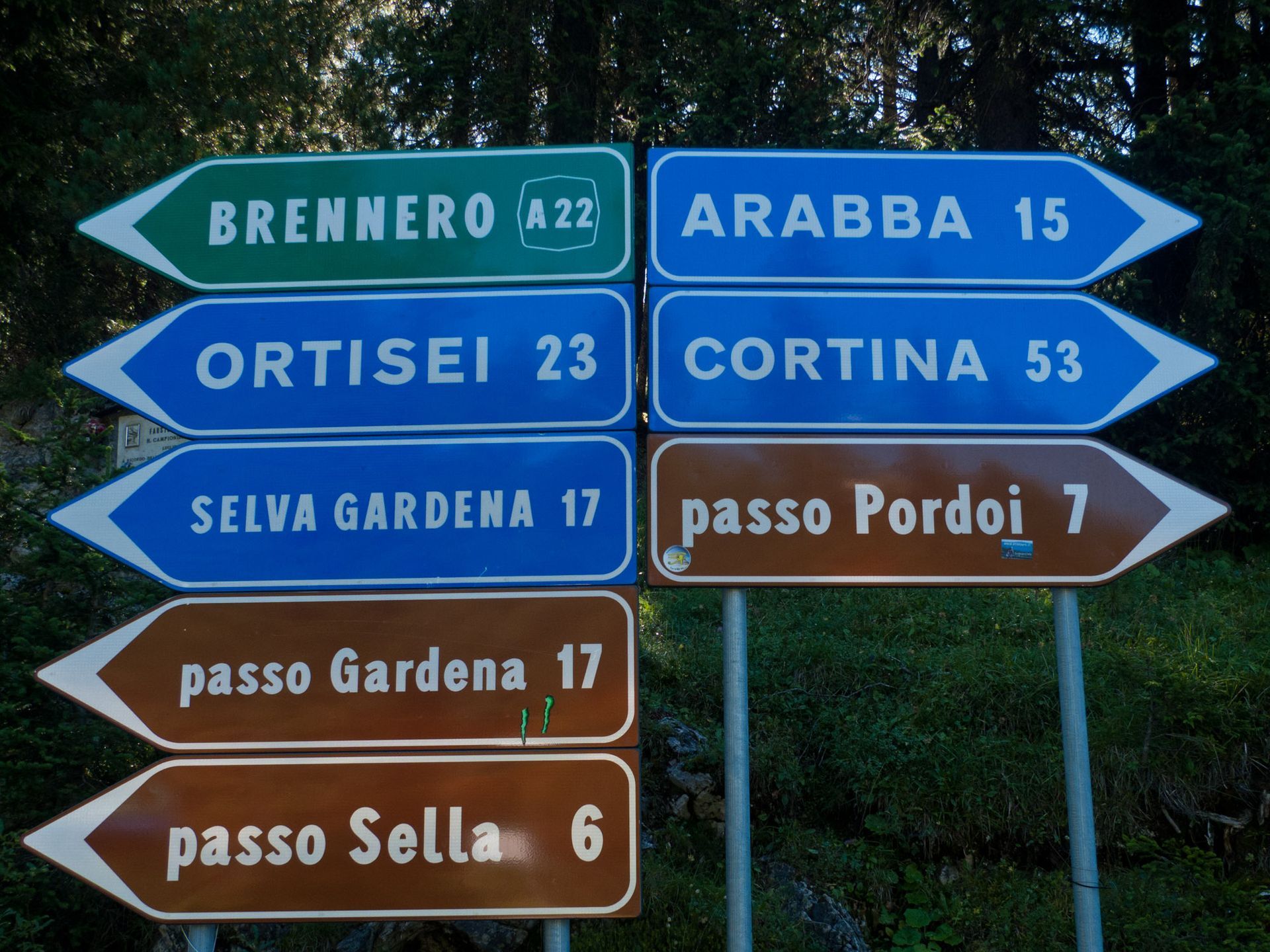 Colorful Alpine trail signs pointing to Brennero, Ortisei, Selva Gardena, Arabba, Cortina, and mountain passes.