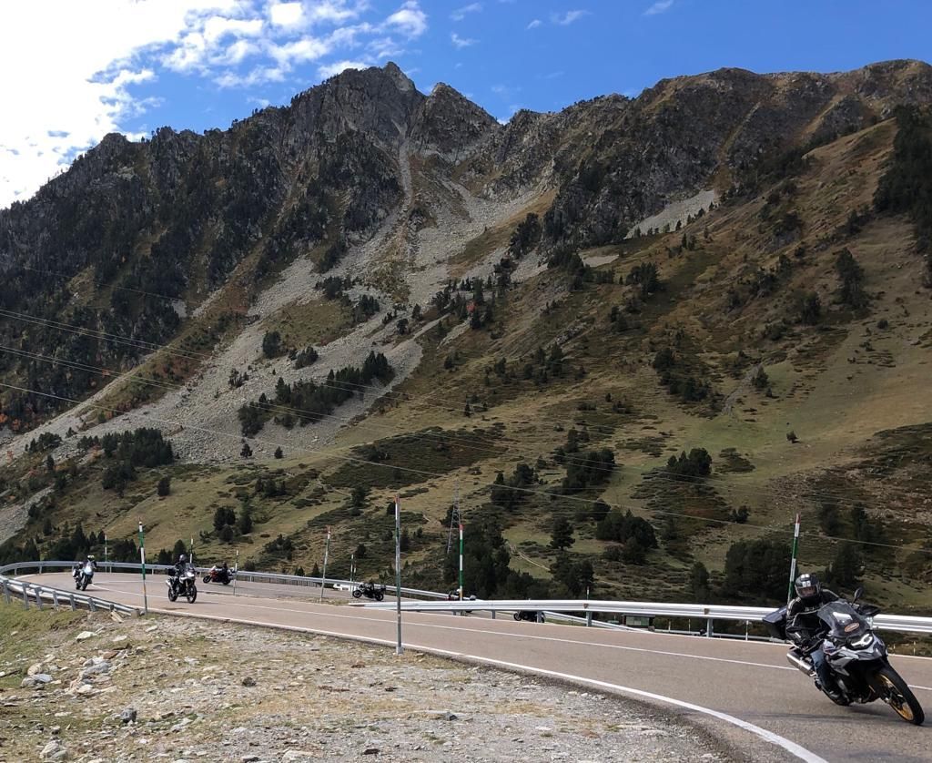 Motorcyclists on a winding mountain road beneath rocky, forested peaks and a blue sky