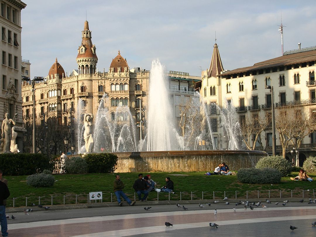 City square with a large fountain, historic buildings, and people sitting on benches in the foreground