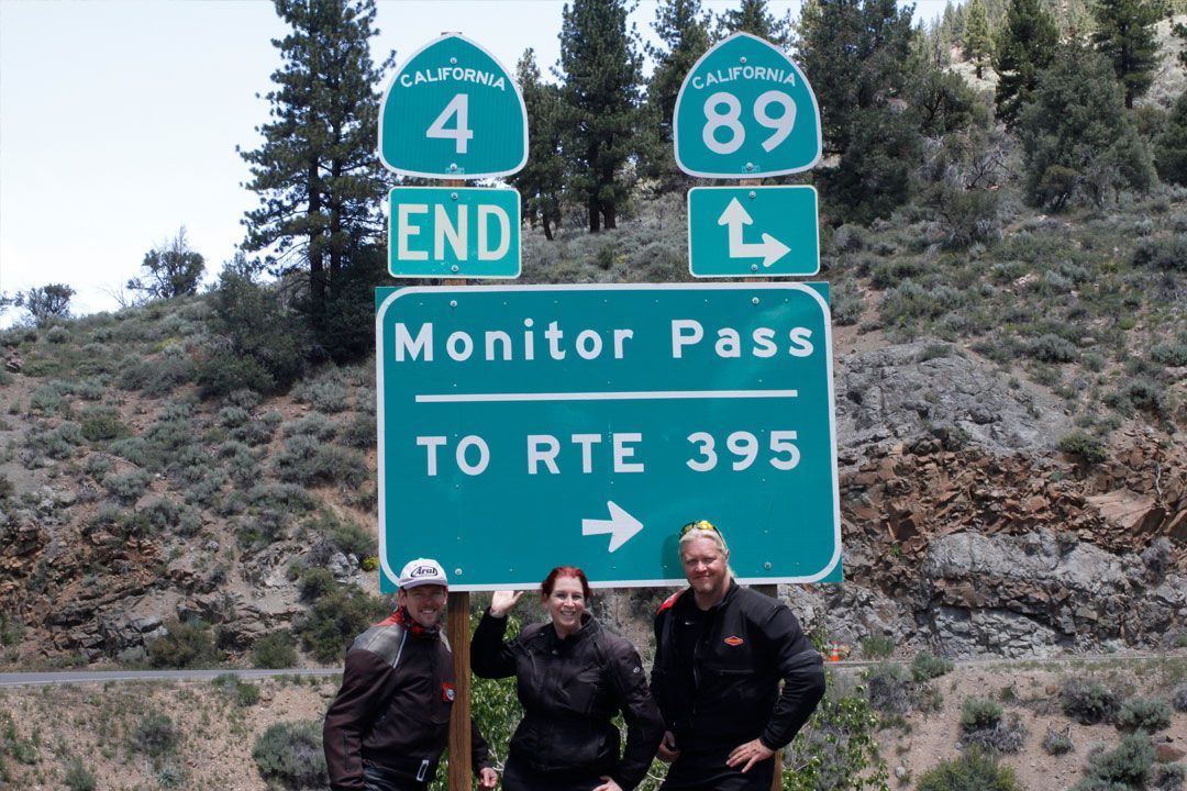 Three hikers posing by Monitor Pass and Route 395 road signs in a rocky mountain landscape