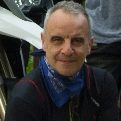 Man in a blue bandana and black jacket, smiling outdoors beside a vehicle