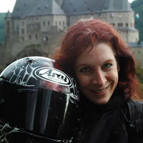 Smiling person with red hair beside a black motorcycle helmet in front of a castle