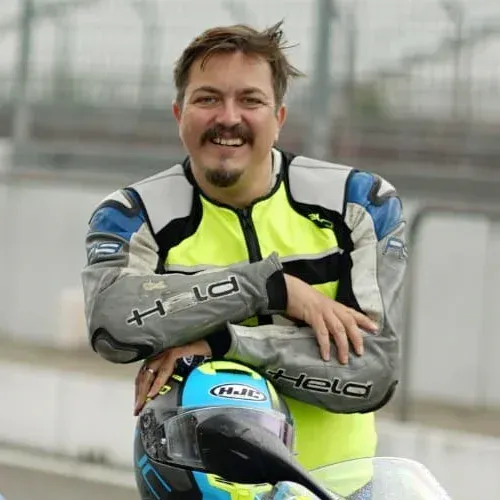 Race car driver in neon yellow and gray suit leaning on a blue race car, smiling at the track.