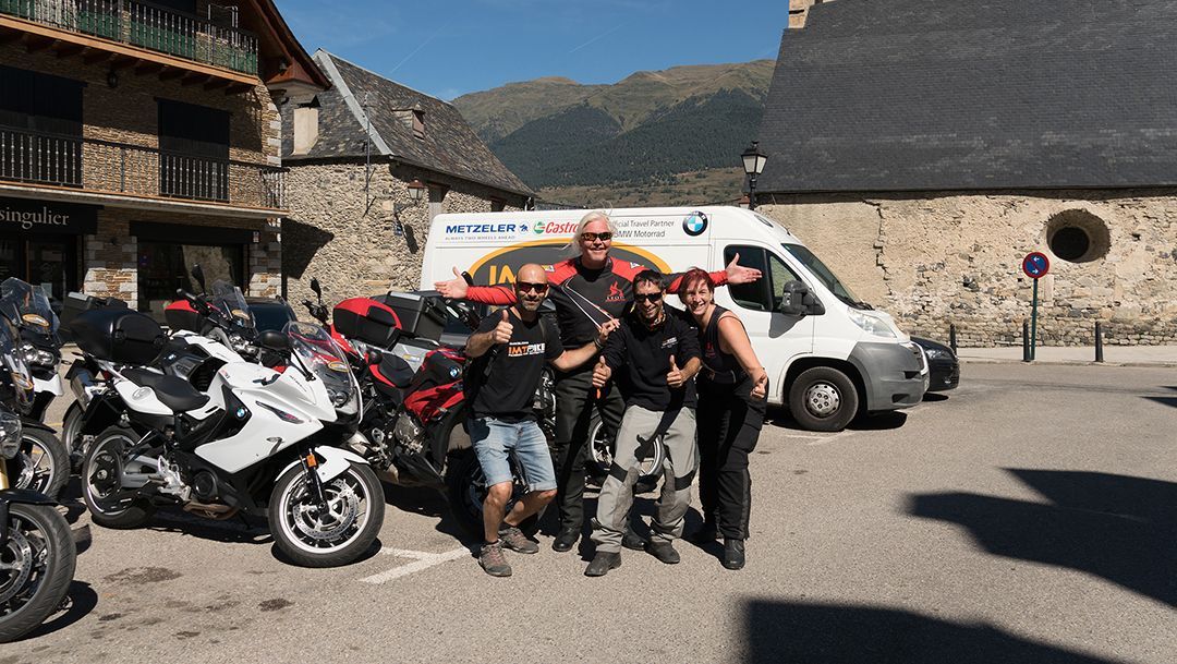 Group of motorcyclists posing beside bikes and a white van in a stone courtyard with mountains behind