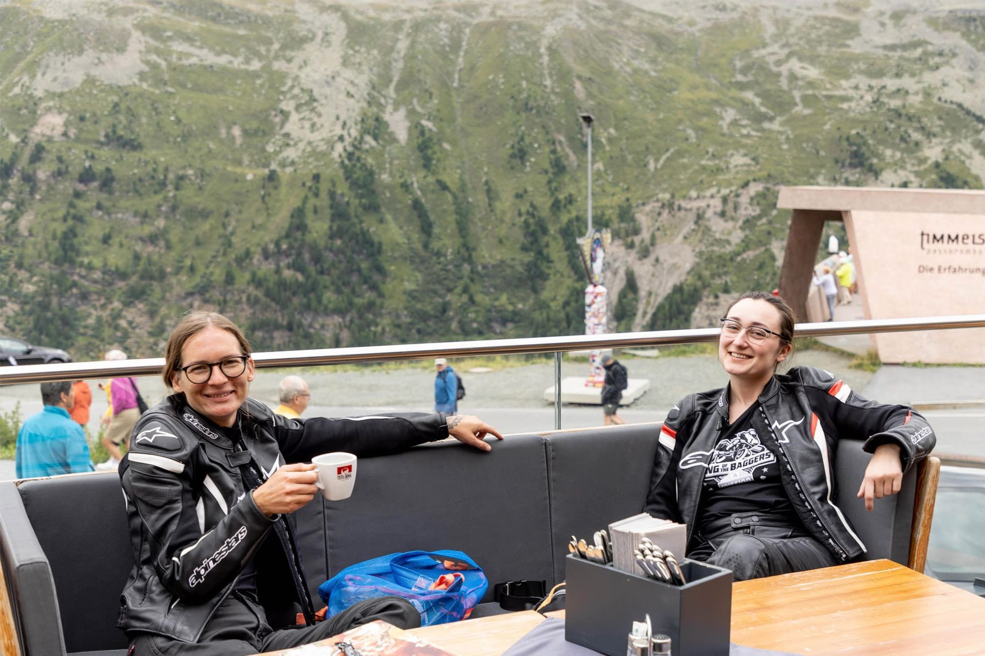 Two people relaxing on a mountain terrace with drinks, overlooking a green valley and cliffs.