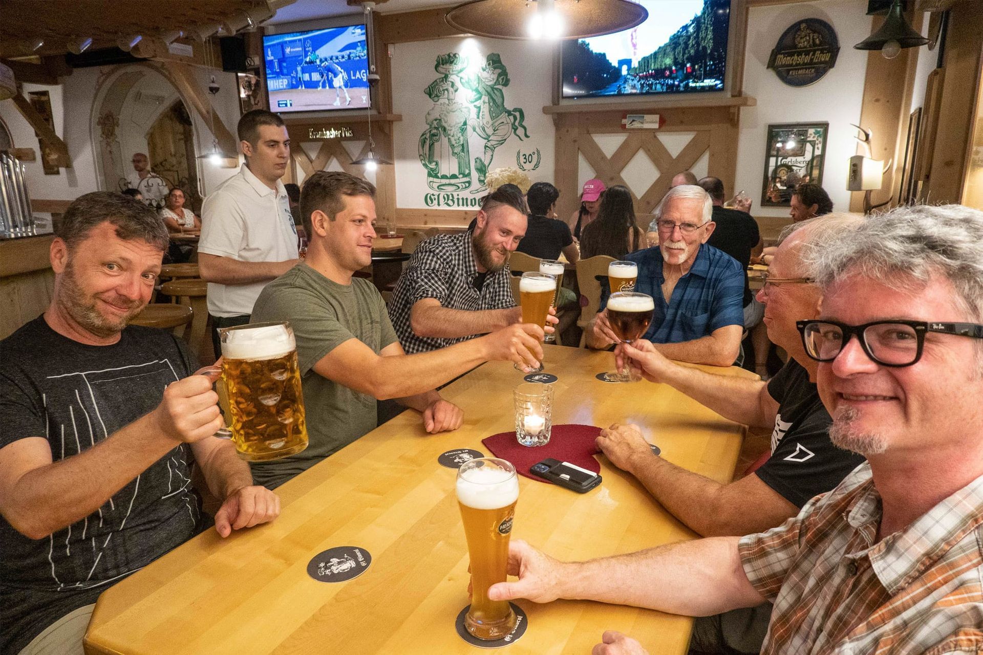 Friends smiling and raising beer mugs at a pub table with TVs in the background