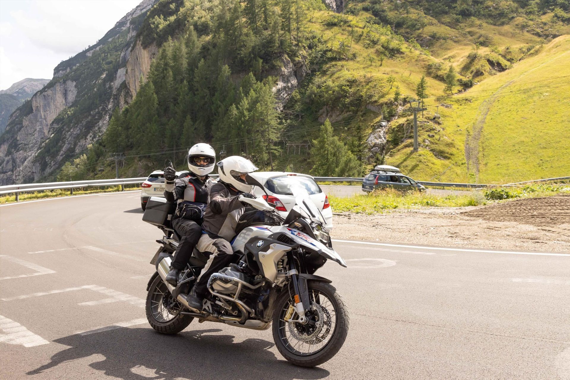 Two motorcyclists on a black bike stopped on a mountain road with green cliffs behind them