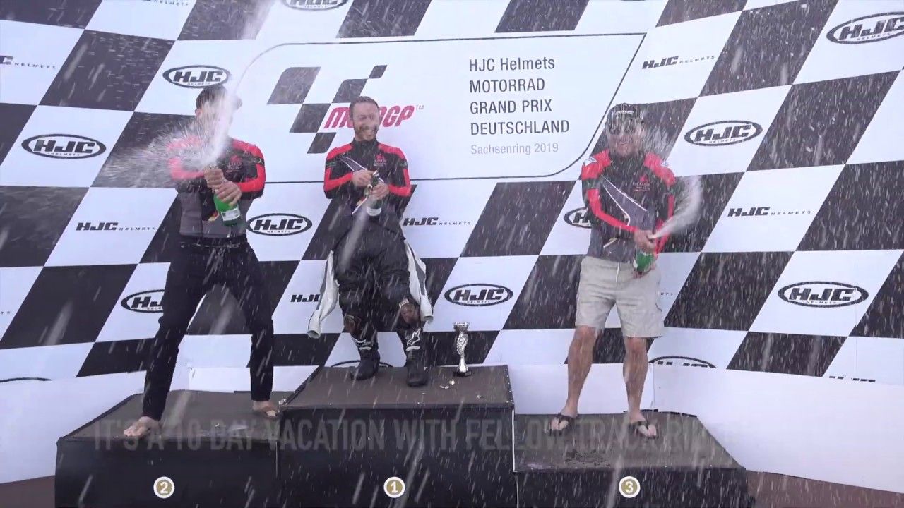 Three people on a podium with champagne spray, holding trophies at a racing event backdrop.