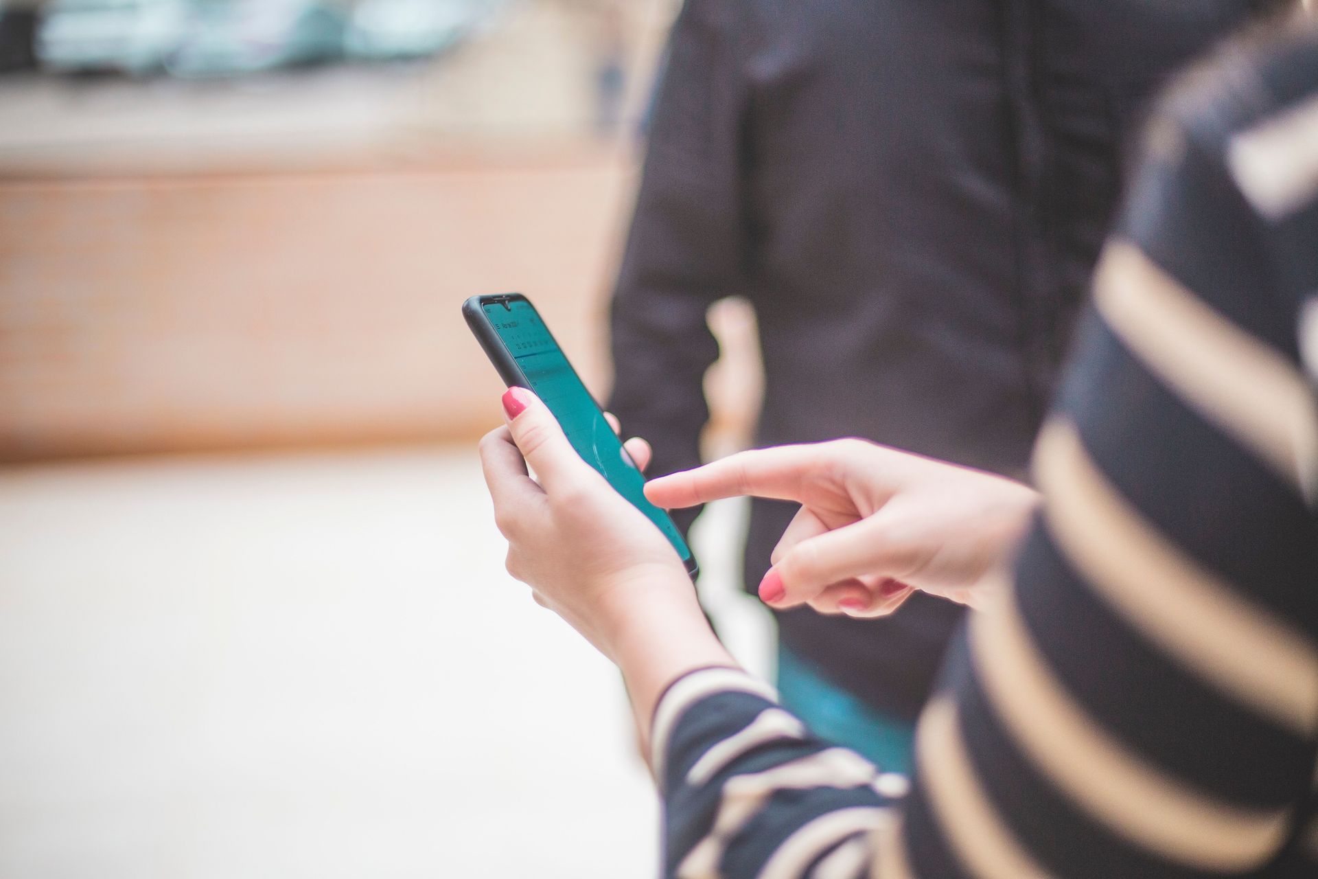 Person using a teal smartphone while seated indoors, with another person blurred in the background