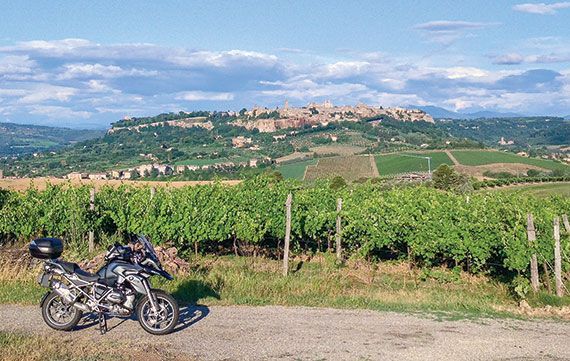 Motorcycle parked beside a vineyard with a hilltop village and blue sky in the distance