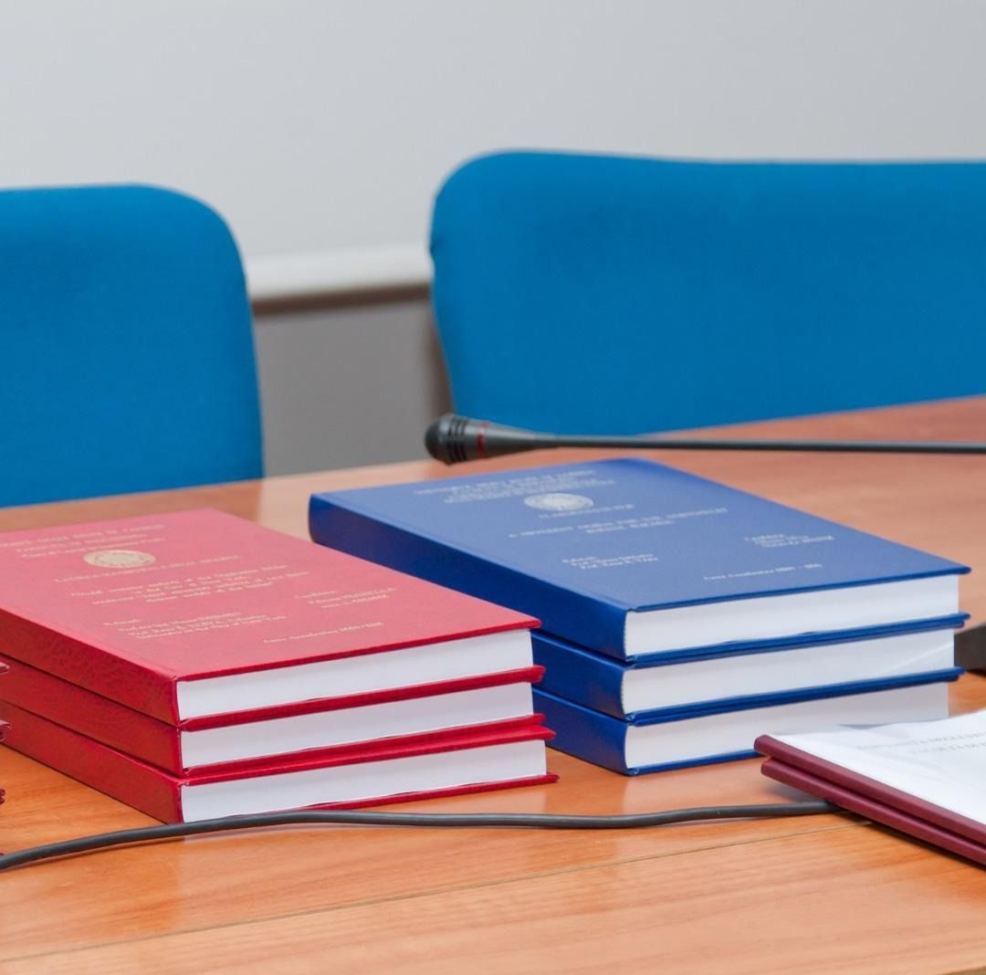 Pile di libri con copertina rigida rossi e blu su un tavolo di legno, con sedie blu sullo sfondo.