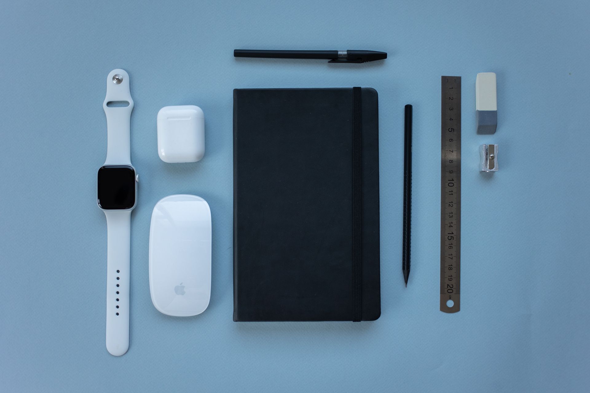Un Apple Watch bianco e degli AirPods sono disposti su una superficie blu insieme a un quaderno nero, una penna, una matita, un righello e una gomma.