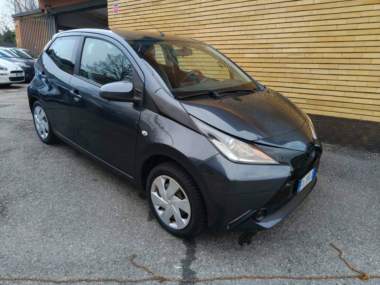 Una Toyota Aygo grigia parcheggiata su un terreno asfaltato di fronte a un edificio in mattoni.
