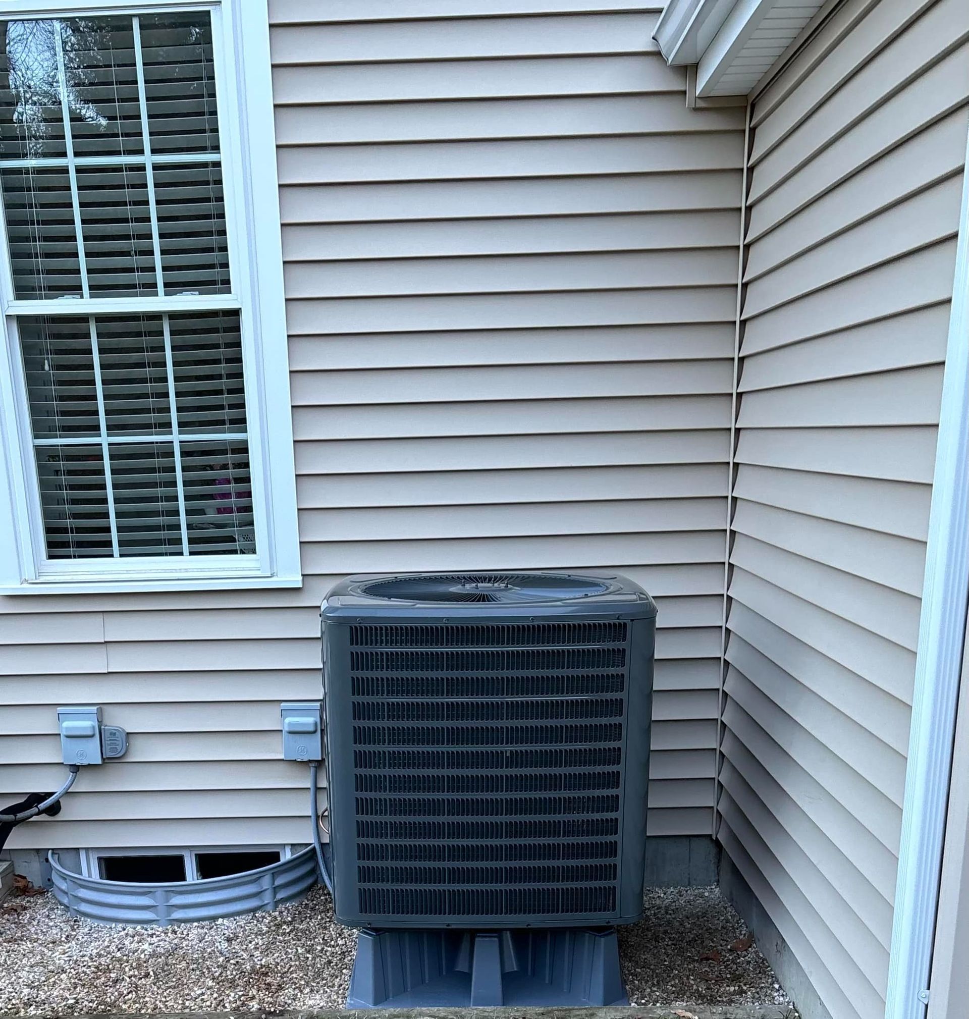Outdoor HVAC unit beside beige house siding near a window and corner trim