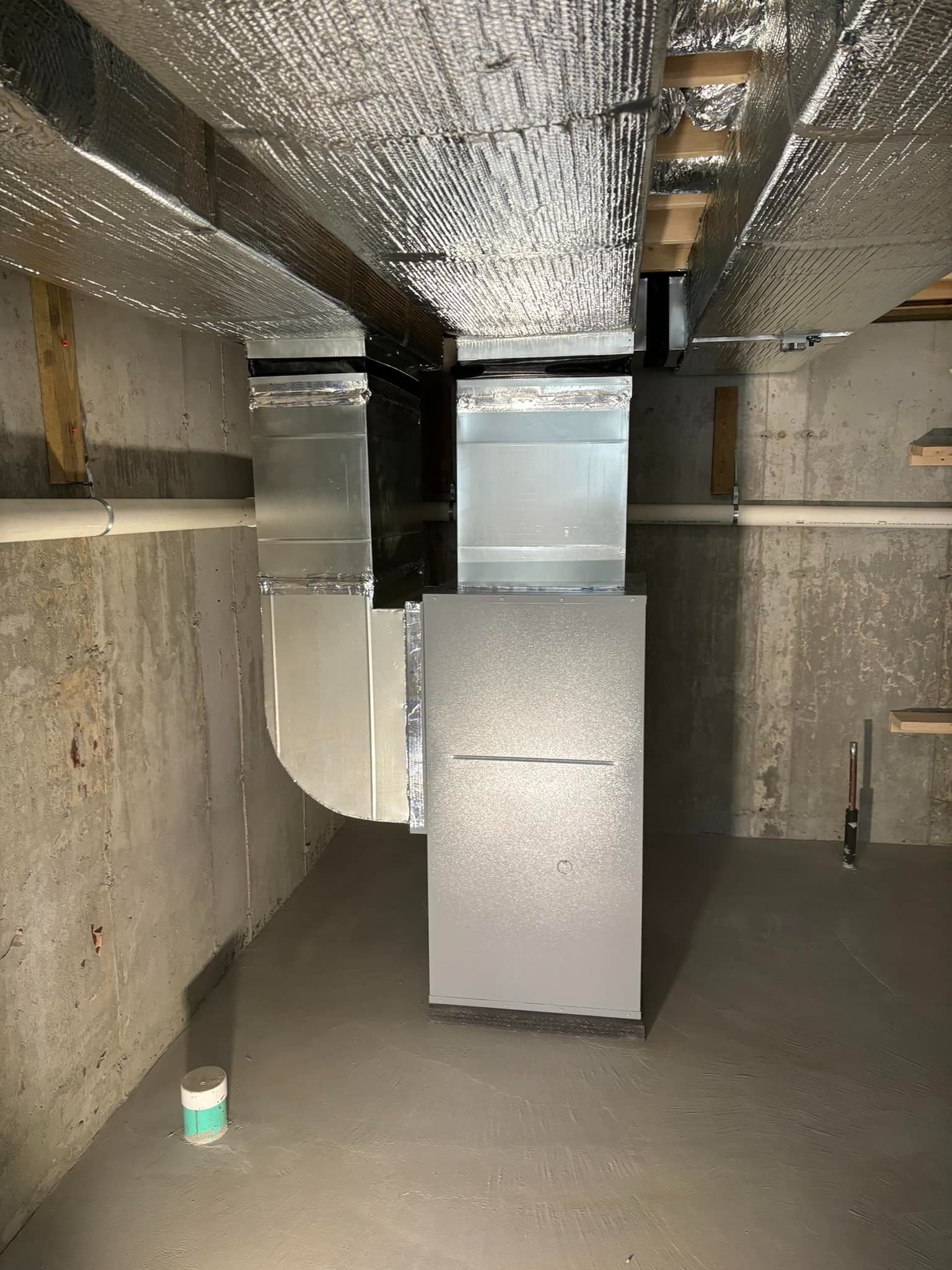 Industrial basement with foil-wrapped ductwork, metal equipment, and concrete walls under exposed ceiling pipes