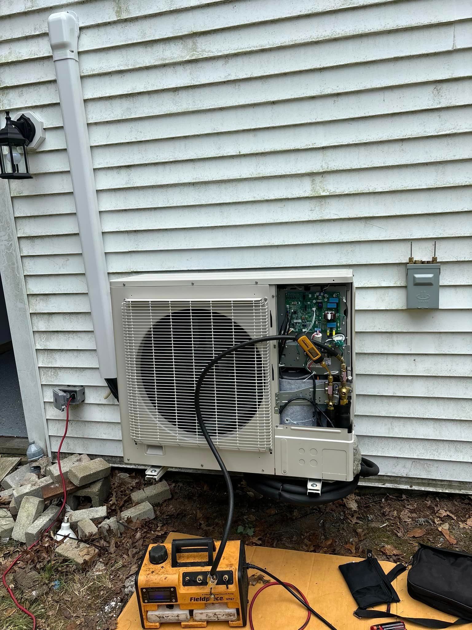 Damaged wall-mounted air conditioner with exposed wiring on white house siding, debris and tools below