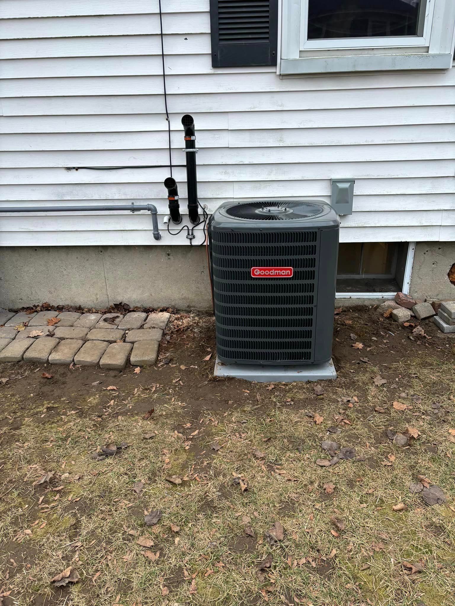 Outdoor HVAC unit beside a white house wall, with exposed pipes and a small gravel patio.