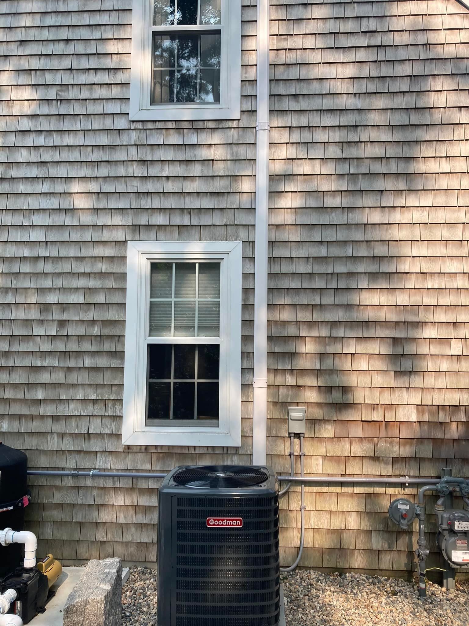 House exterior with shingle siding, two windows, an air conditioner, and a black trash bin below.