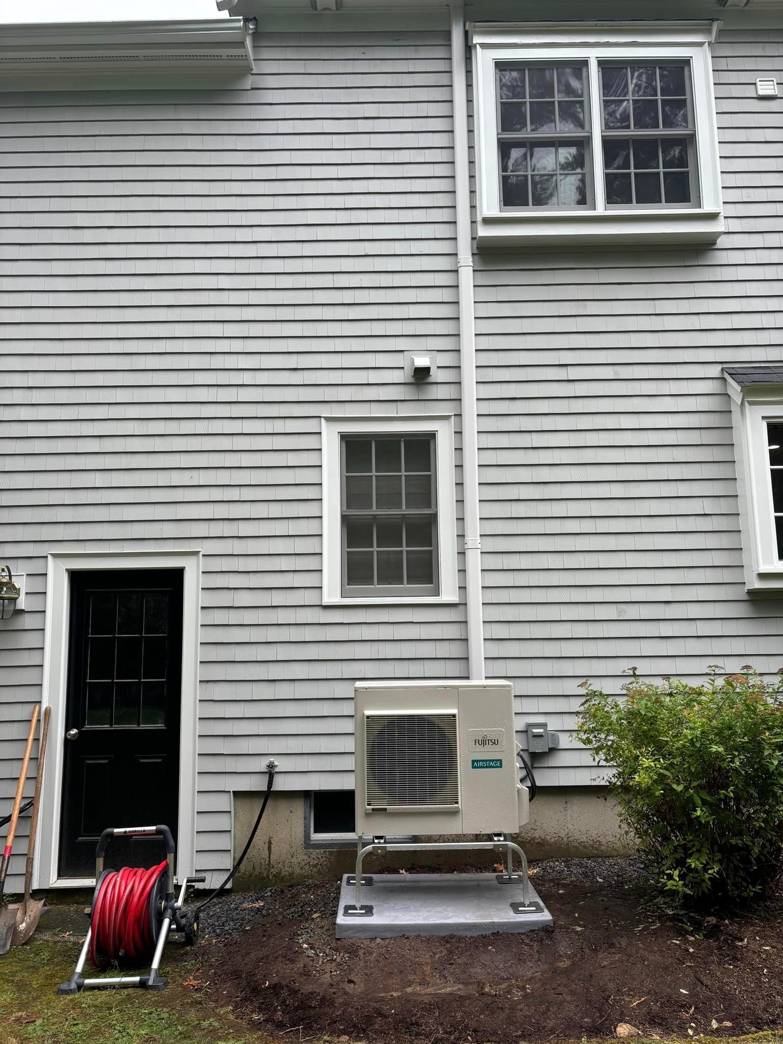 Back of a gray house with white-trim windows, utility boxes, a hose reel, and shrubs by the wall