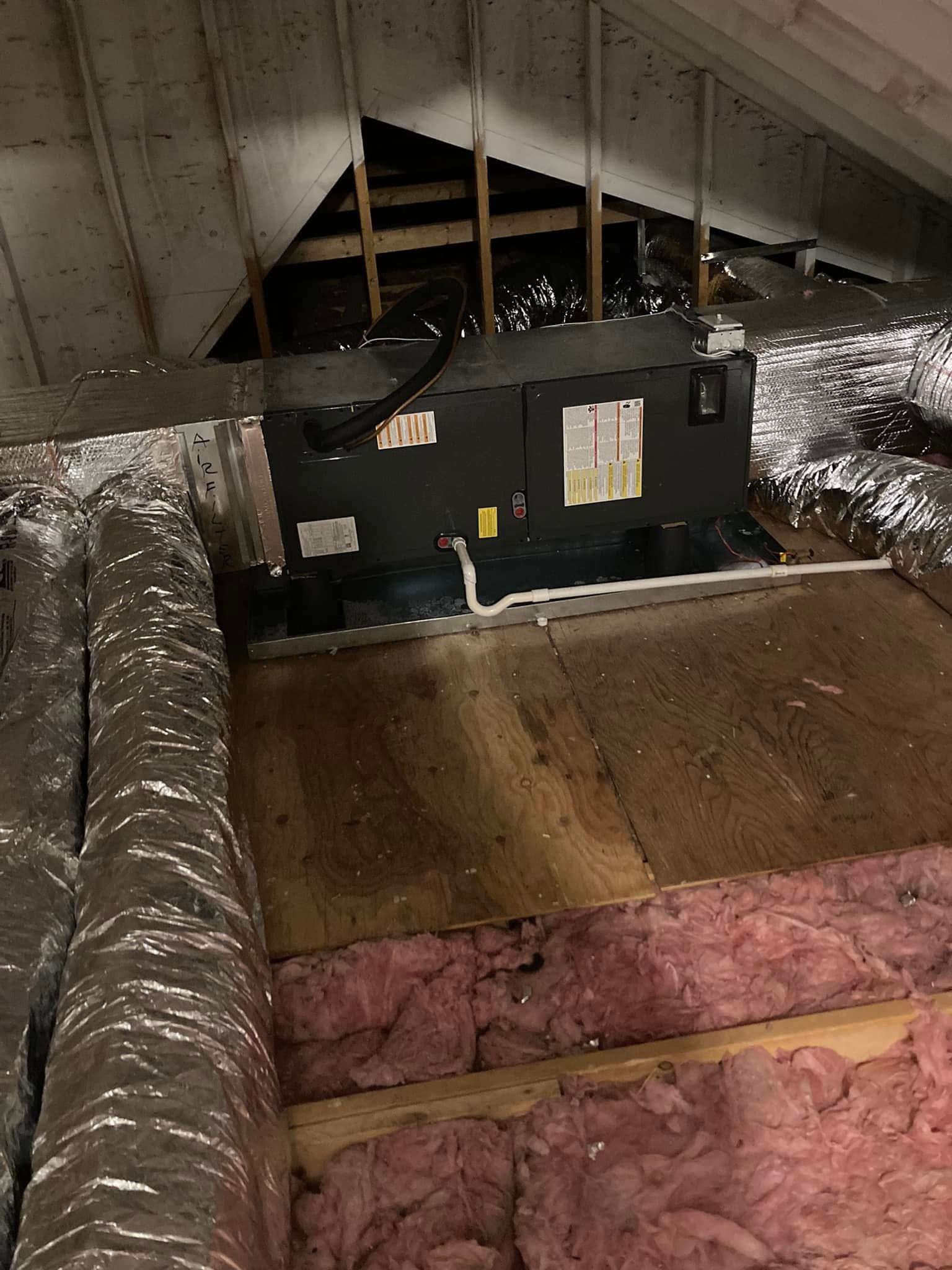 Attic HVAC unit amid silver ductwork and pink insulation under sloped roof rafters