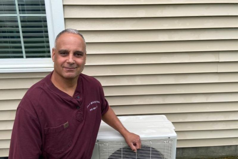 Man standing beside a white outdoor air conditioning unit next to a beige house siding wall