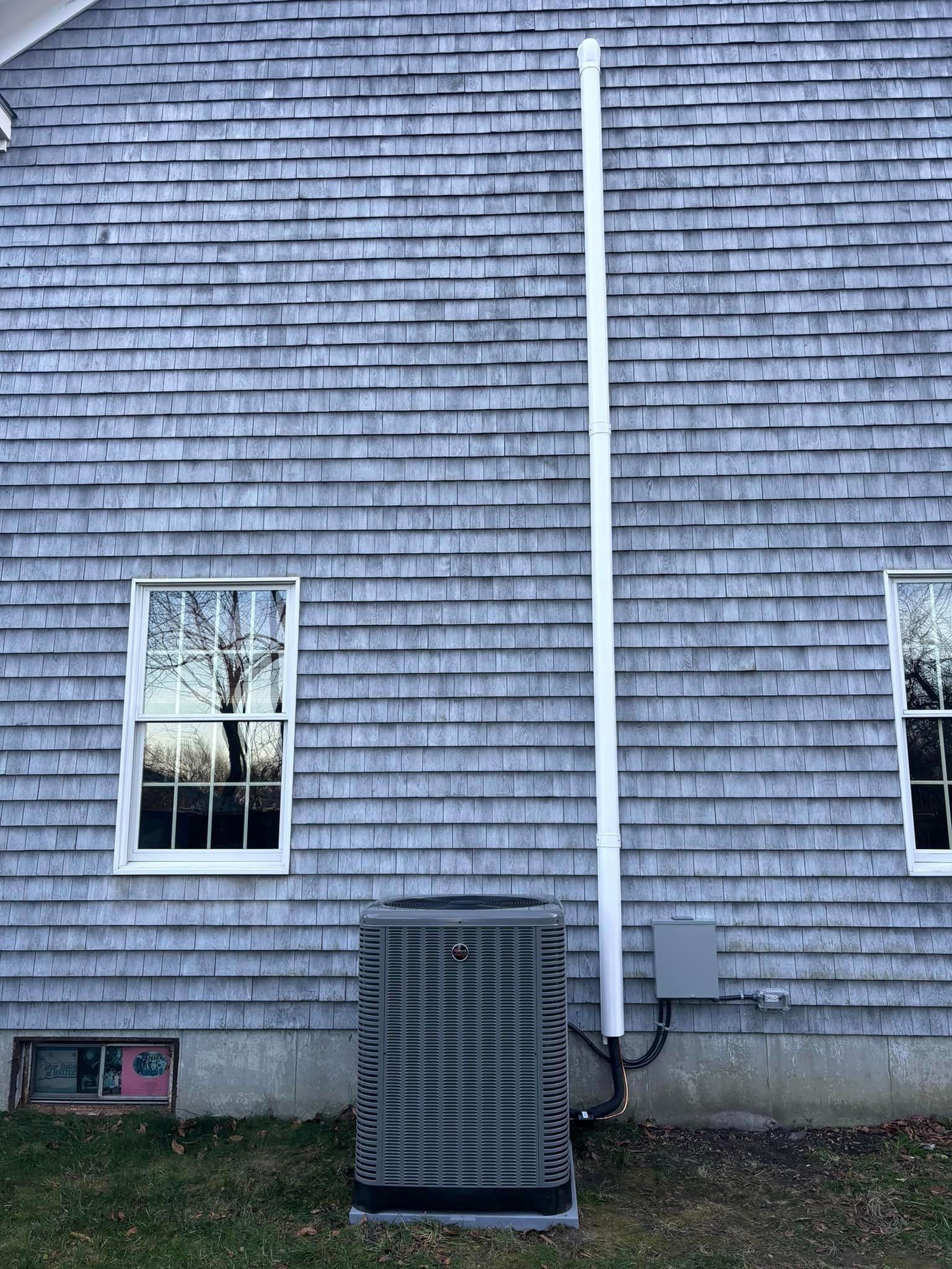 Gray shingled house wall with two windows, a white downspout, and an air conditioning unit below.