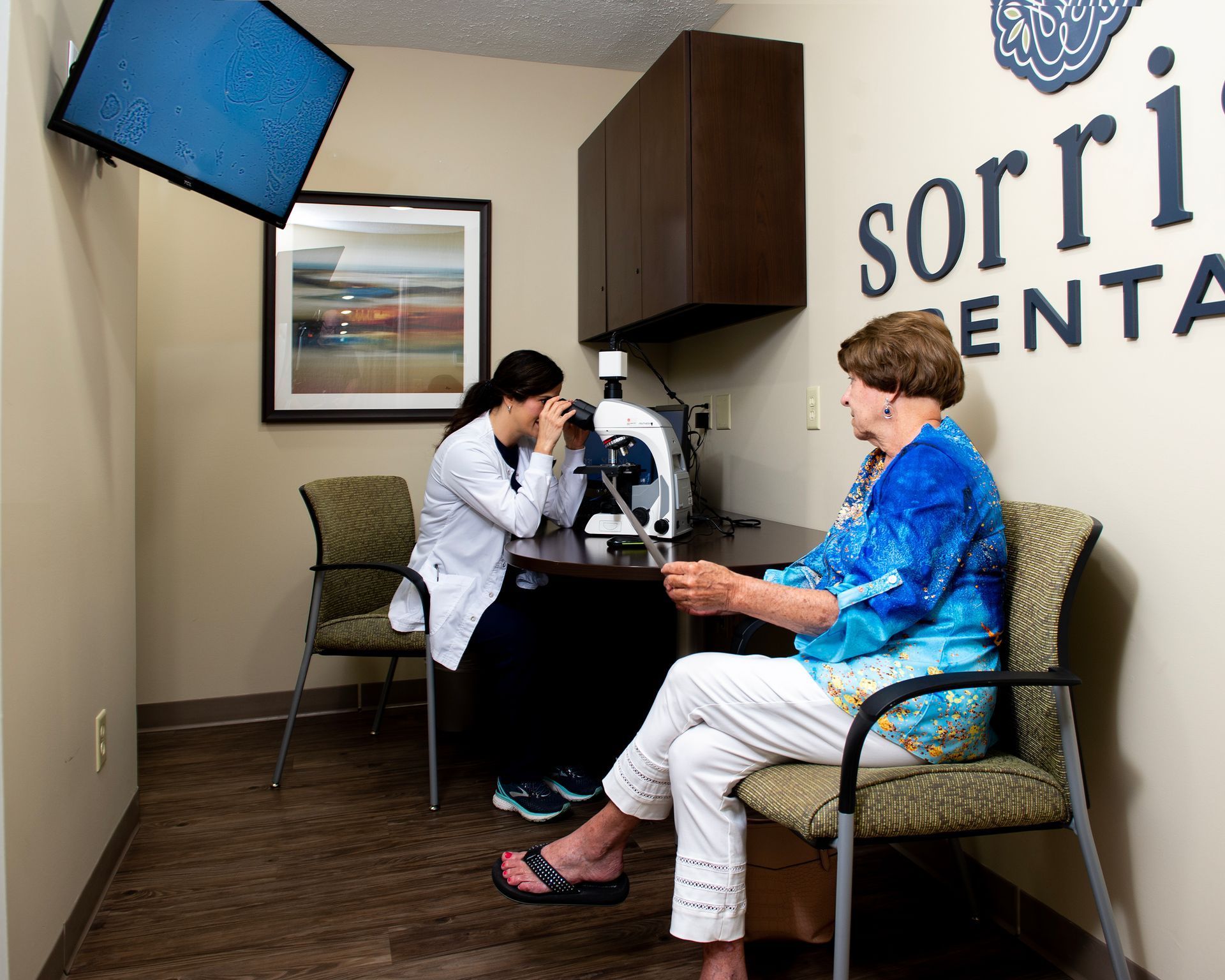 A woman is sitting in a chair while a nurse looks through a microscope.