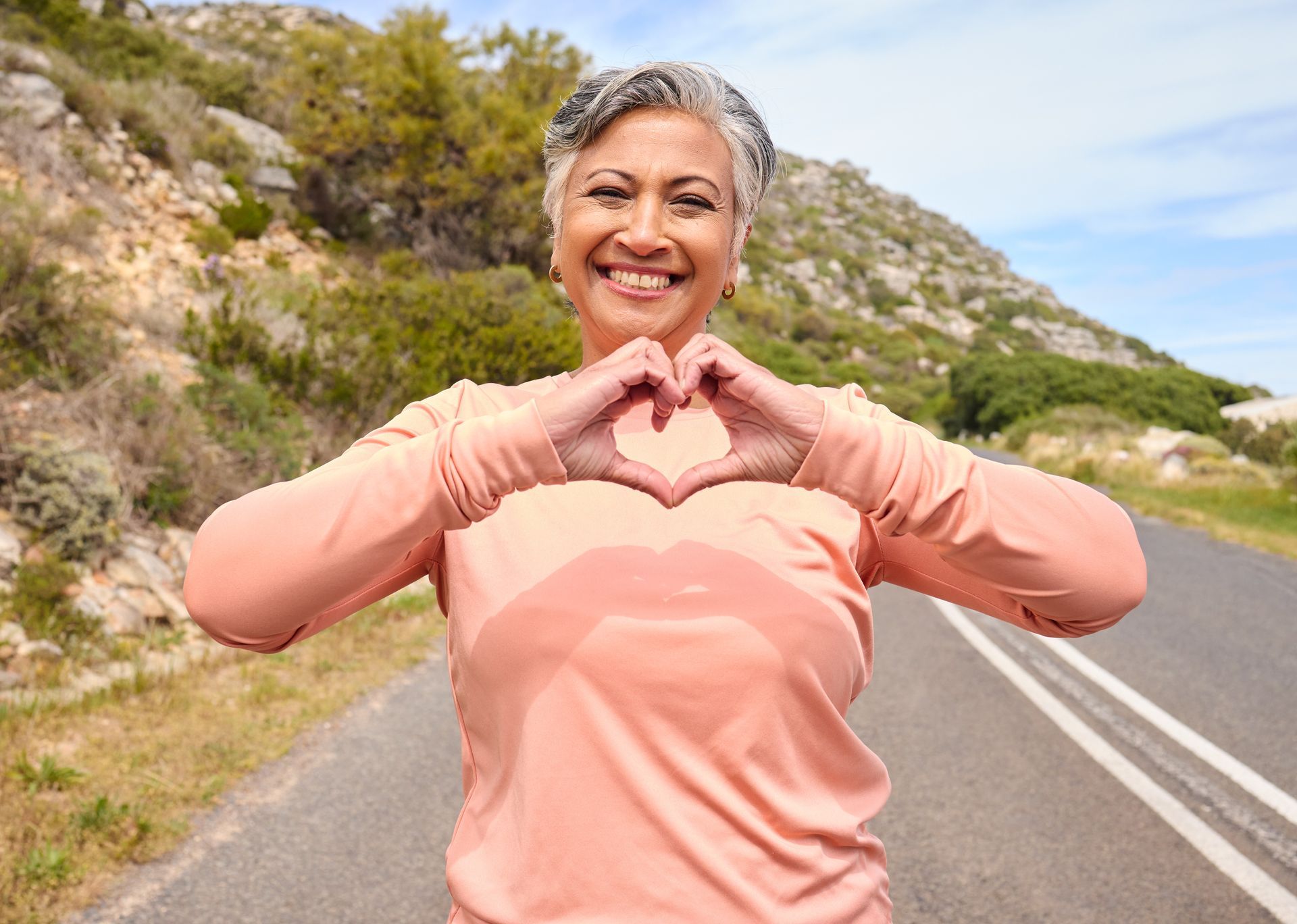 Smiling person in a pink shirt making a heart shape with their hands on a roadside