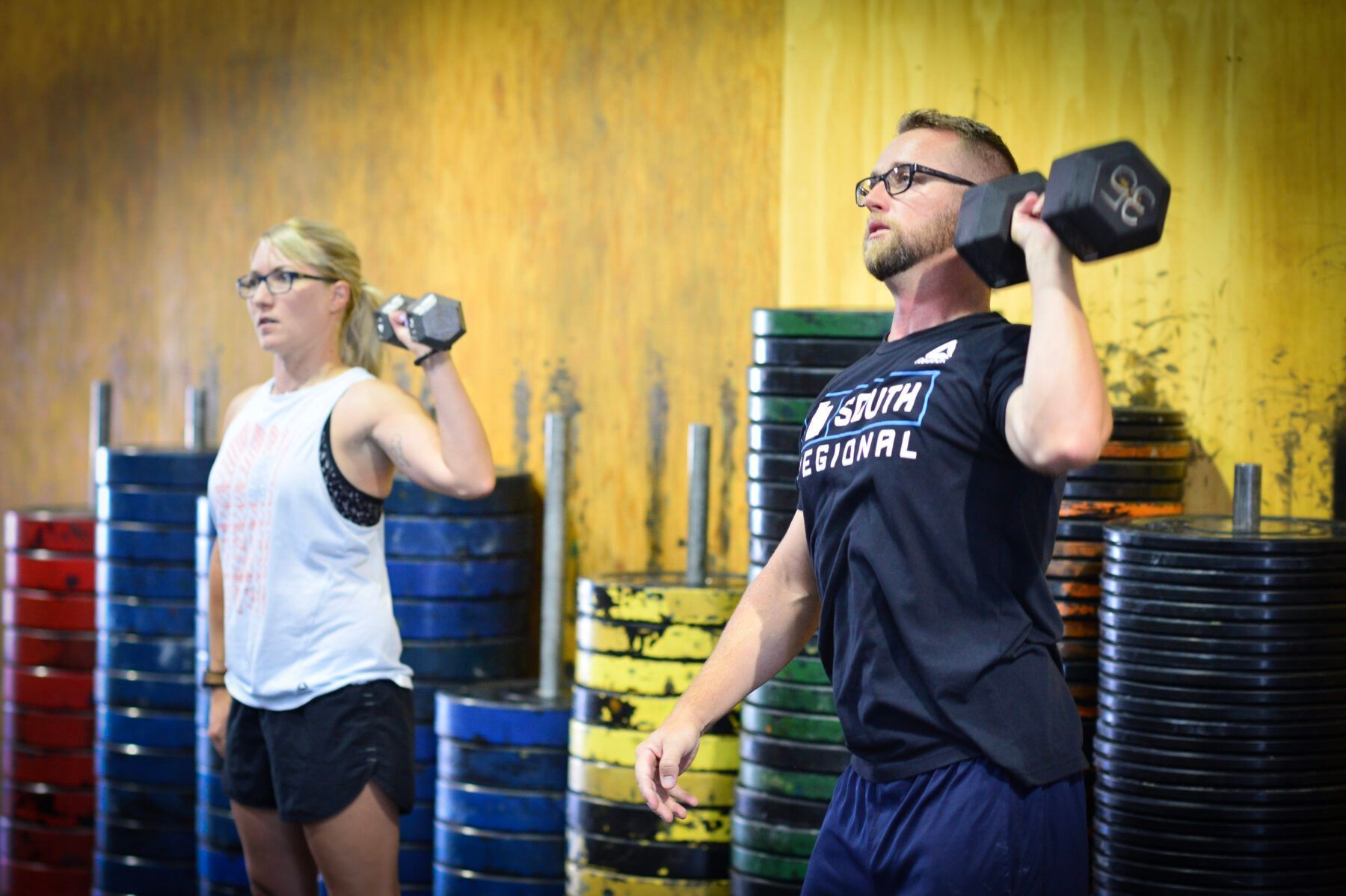 Two people lifting dumbbells in Iron Grip CrossFit, a gym in Tucson, AZ, standing in front of stacked weights and a yellow wall.