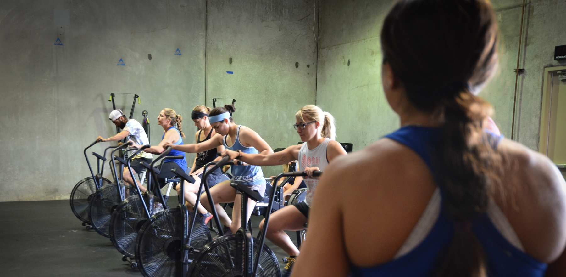 People exercising on stationary bikes in Iron Grip CrossFit, a gym in Tucson, AZ, viewed past a person in the foreground