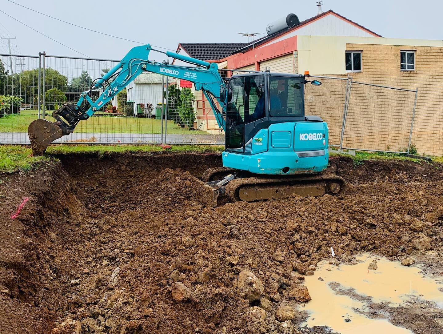Blue excavator digging in muddy ground next to a building. — Lifestyle Demolition In Byron Bay, NSW