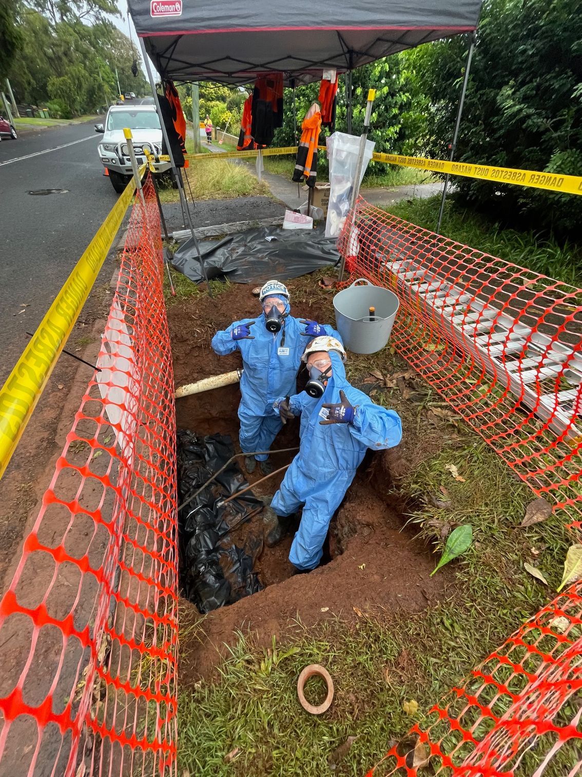 Two People in Blue Protective Hazmat Suits and Respirators Pose Inside a Dug-Out Trench Surrounded by Orange Safety Mesh — Lifestyle Demolition In Murwillumbah, NSW