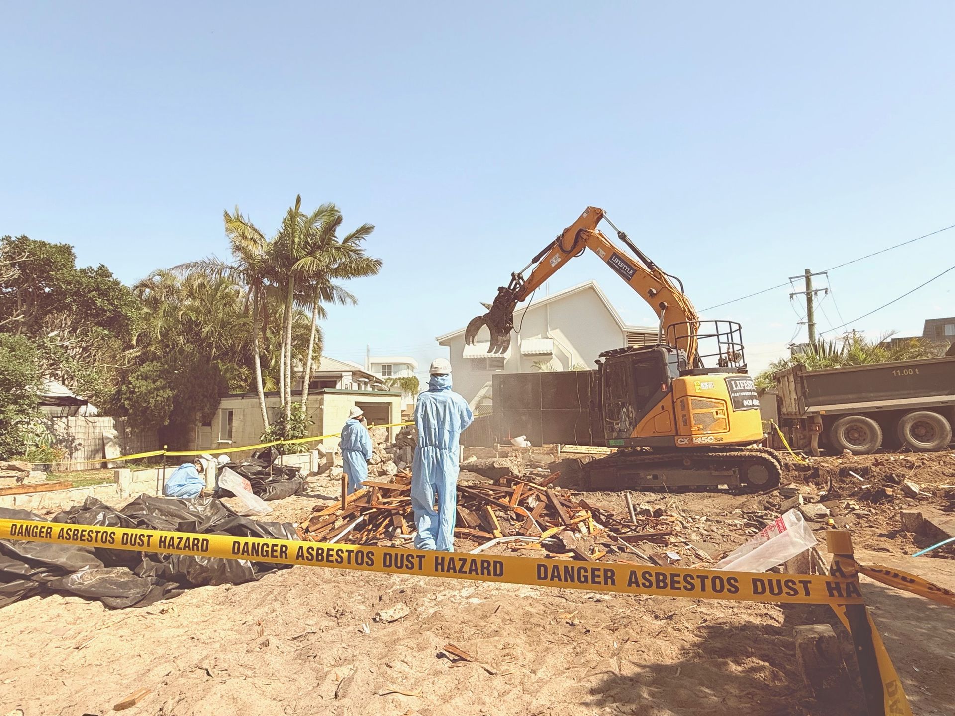 Workers in protective suits at an asbestos demolition site with an excavator, truck, and yellow warning tape — Lifestyle Demolition In Mullumbimby, NSW