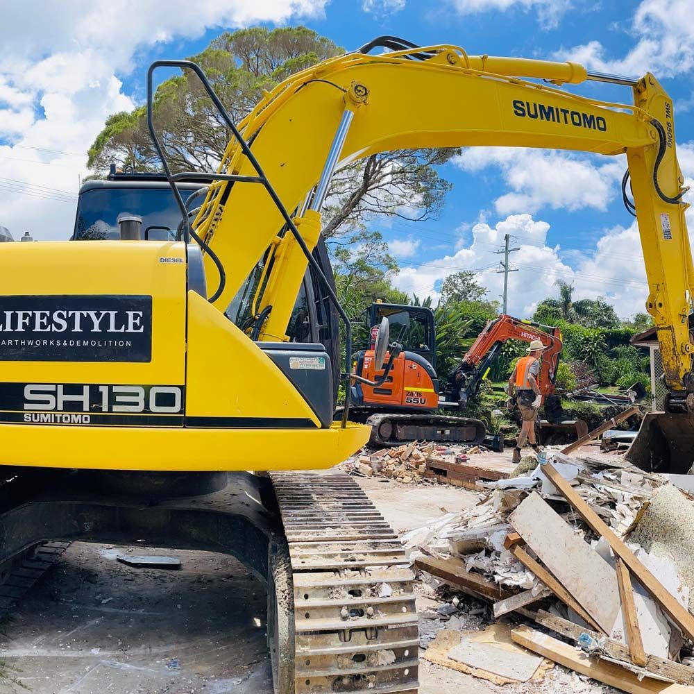 A yellow Sumitomo excavator parked at a construction site with debris in the foreground and a smaller orange excavator — Lifestyle Demolition in Kingscliff, NSW