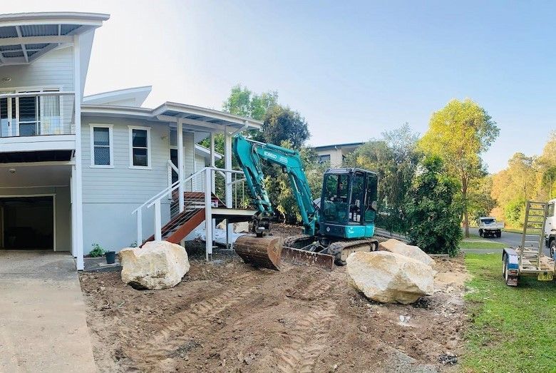 A Small Excavator Digs in Front of a Light-colored House, With Large Boulders Nearby — Lifestyle Demolition In Byron Bay, NSW