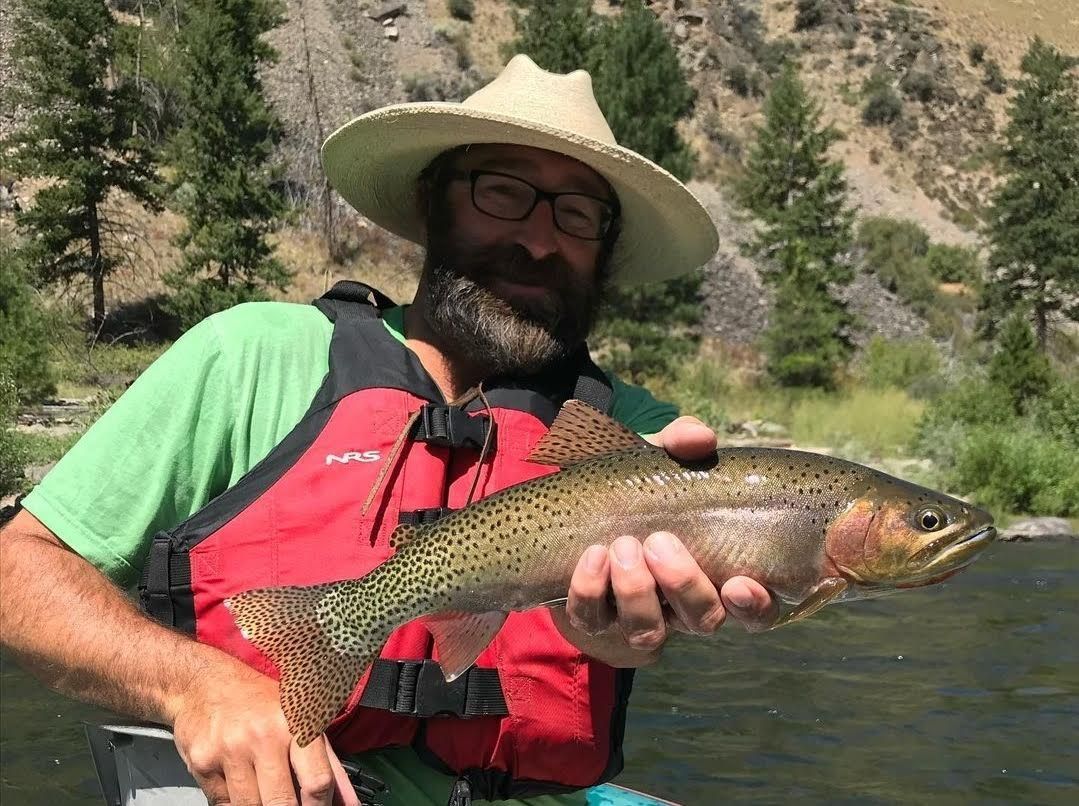 A man in a hat is holding a rainbow trout in his hands.
