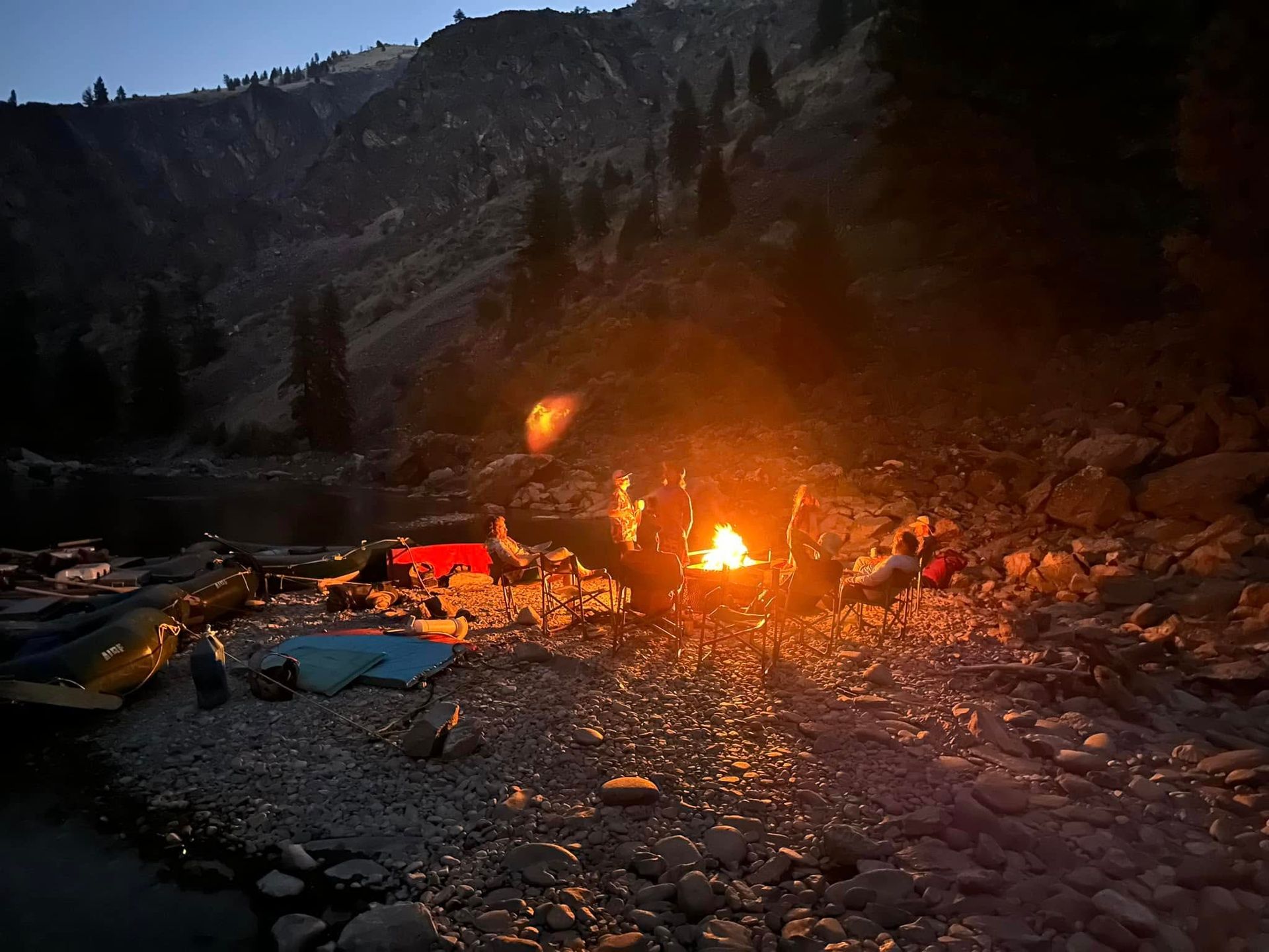 A group of people are sitting around a campfire on a rocky beach.
