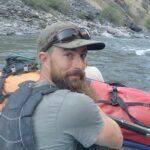 A man with a beard is sitting in a boat on a river.