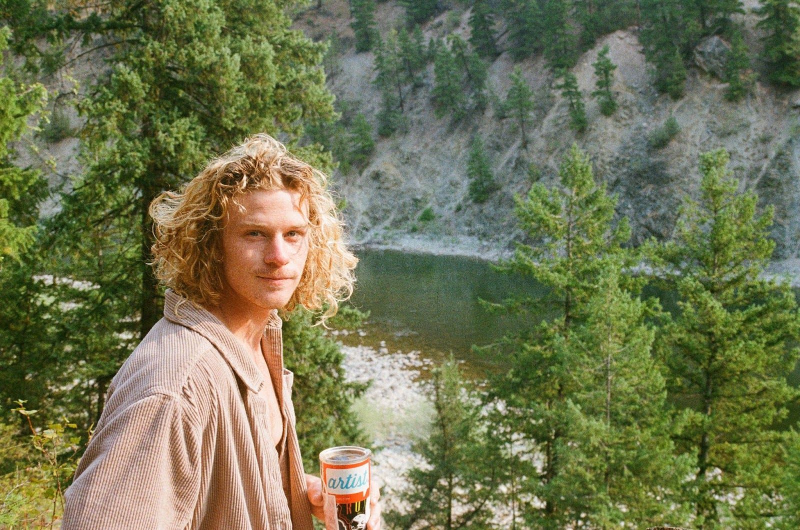 A young man with curly hair is standing in front of a lake holding a can of soda.