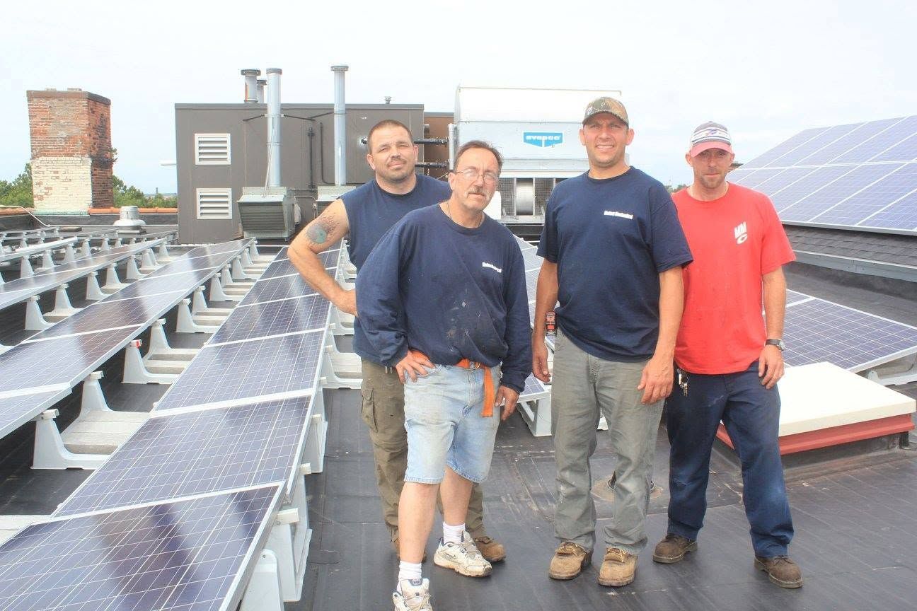Three people standing on a rooftop beside rows of solar panels and industrial equipment