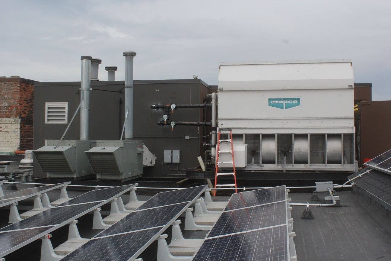 Rooftop HVAC units and solar panels on a flat commercial roof, with a ladder leaning against a wall.