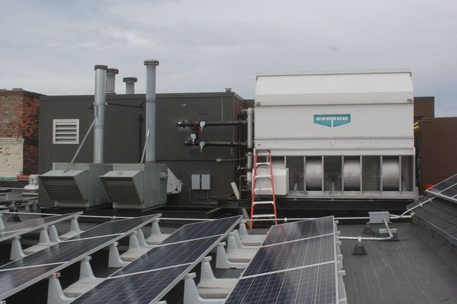Rooftop with HVAC units, vents, solar panels, and a ladder beside a large white rooftop structure