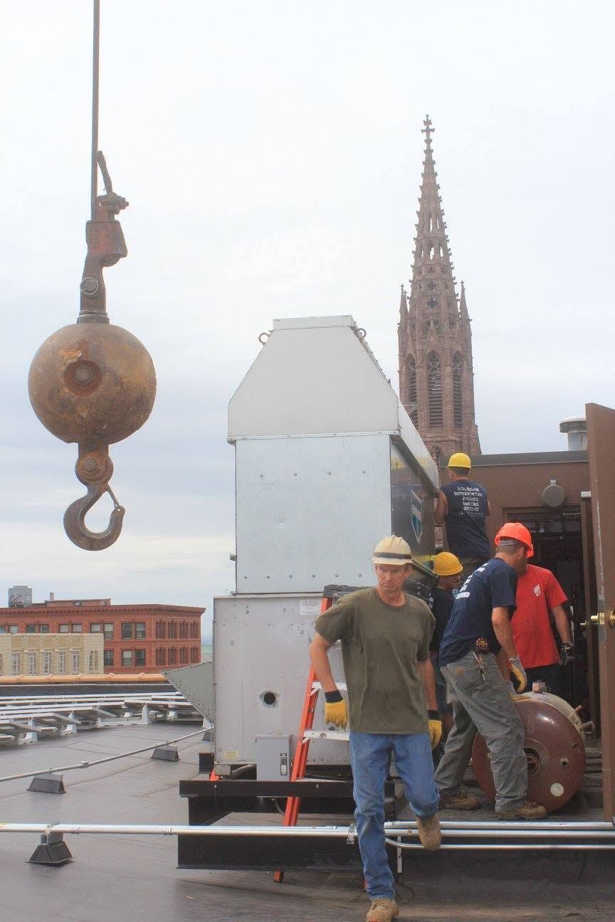 Workers on a rooftop with a crane hook and a tall brick tower in the background.
