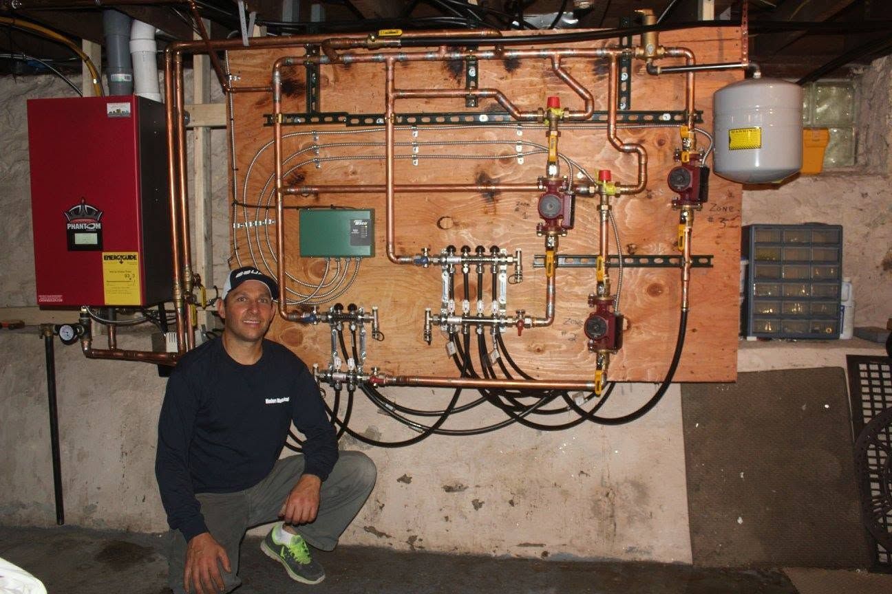 Man kneeling beside a wall of pipes, valves, and gauges in a basement mechanical room