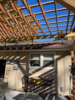 Wooden pergola built over a house's back patio. The sky is visible through the rafters.
