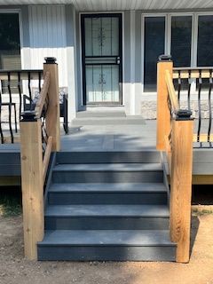 Gray outdoor stairs with wooden posts and black railings lead to a house with a screen door.